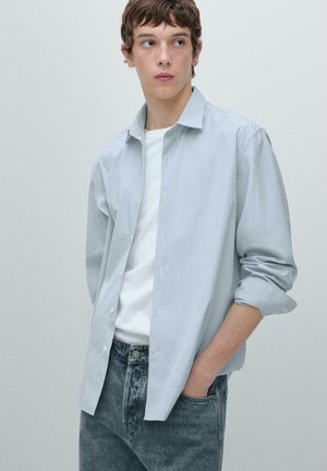 Jeune homme aux cheveux courts bruns portant une chemise bleu clair à fines rayures sur un t-shirt blanc et un jean noir délavé, main dans la poche, regardant sur le côté.