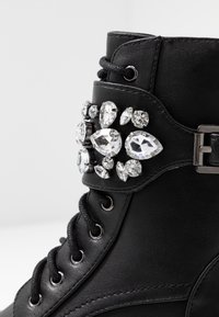 Černá kožená bota s kulatou špičkou, vybavená shlukem různě tvarovaných průhledných rhinestones na pásku kolem kotníku a černými šňůrkami.