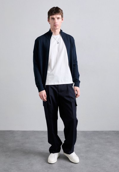 Temno modra jakna na zadrgo čez belo majico, kombinirana s temnimi cargo hlačami in belimi športnimi copati. Preprost dizajn s sproščenim krojem.