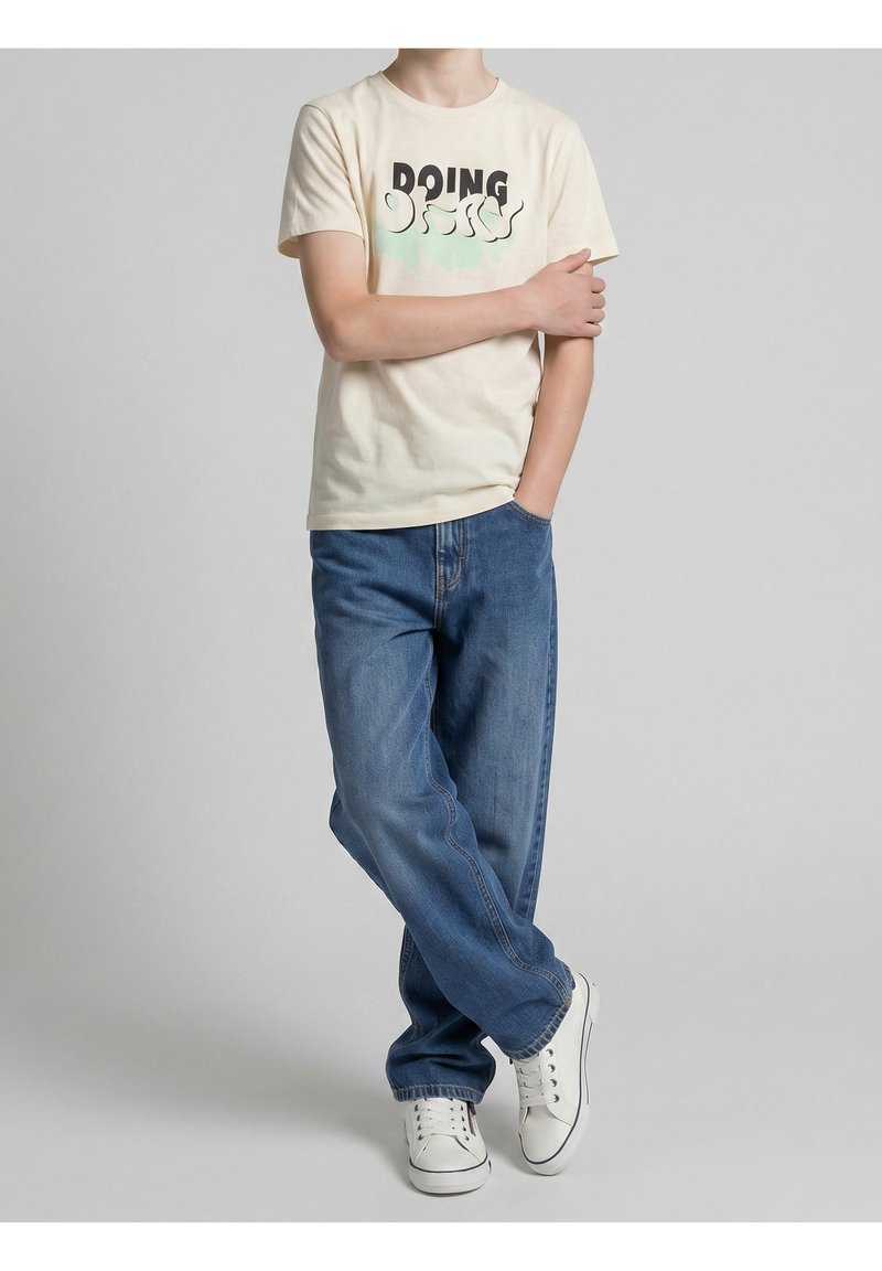 Personne portant un t-shirt crème avec le texte "DOING", jean bleu, baskets blanches, debout avec une jambe croisée et une main sur le bras opposé.