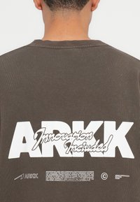 Dark brown t-shirt featuring a large white logo with "ARKK" and "Innovation Included" in stylized text on the back. Ribbed collar.