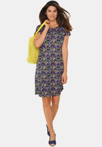 Blumendruckkleid mit kurzen Ärmeln in Marineblau, das bunte Blattformen zeigt. Getragen mit einer gelben geflochtenen Tasche und blauen Keilabsätzen.