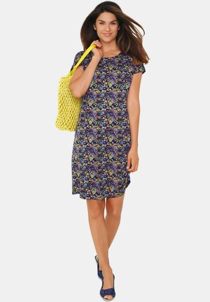 Blumendruckkleid mit kurzen Ärmeln in Marineblau, das bunte Blattformen zeigt. Getragen mit einer gelben geflochtenen Tasche und blauen Keilabsätzen.
