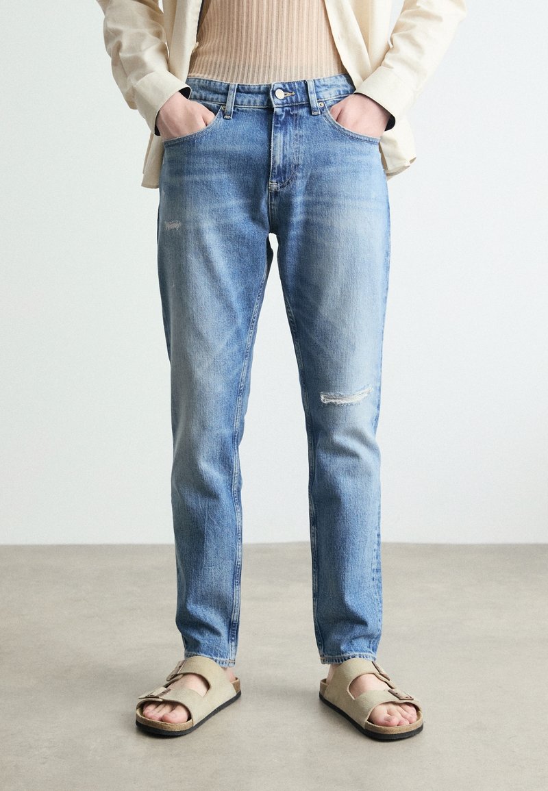 Ljusblå denimjeans med avslappnad passform, med subtil slitning och knäppning. Bärs med beige sandaler.