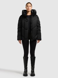 Schwarze Steppjacke mit Kapuze, mit einem gesteppten Design und Reißverschluss. Kombiniert mit schwarzen Leggings und kniehohen schwarzen Stiefeln.