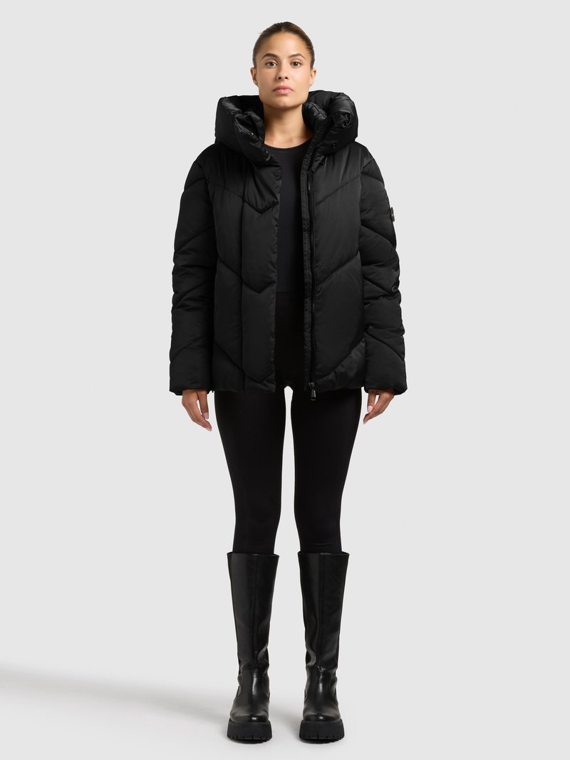 Schwarze Steppjacke mit Kapuze, mit einem gesteppten Design und Reißverschluss. Kombiniert mit schwarzen Leggings und kniehohen schwarzen Stiefeln.