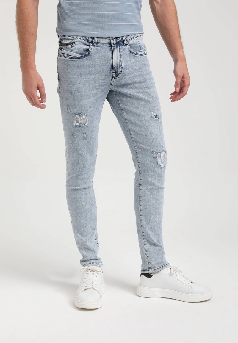 Homme portant un jean skinny bleu clair déchiré avec des patches et des baskets blanches, debout devant un fond uni.