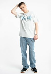 Młody mężczyzna w jasnoniebieskiej koszulce z grafiką, luźnych niebieskich dżinsach z łańcuchem, czarnych sneakersach i zegarku, pozujący z ręką na głowie.