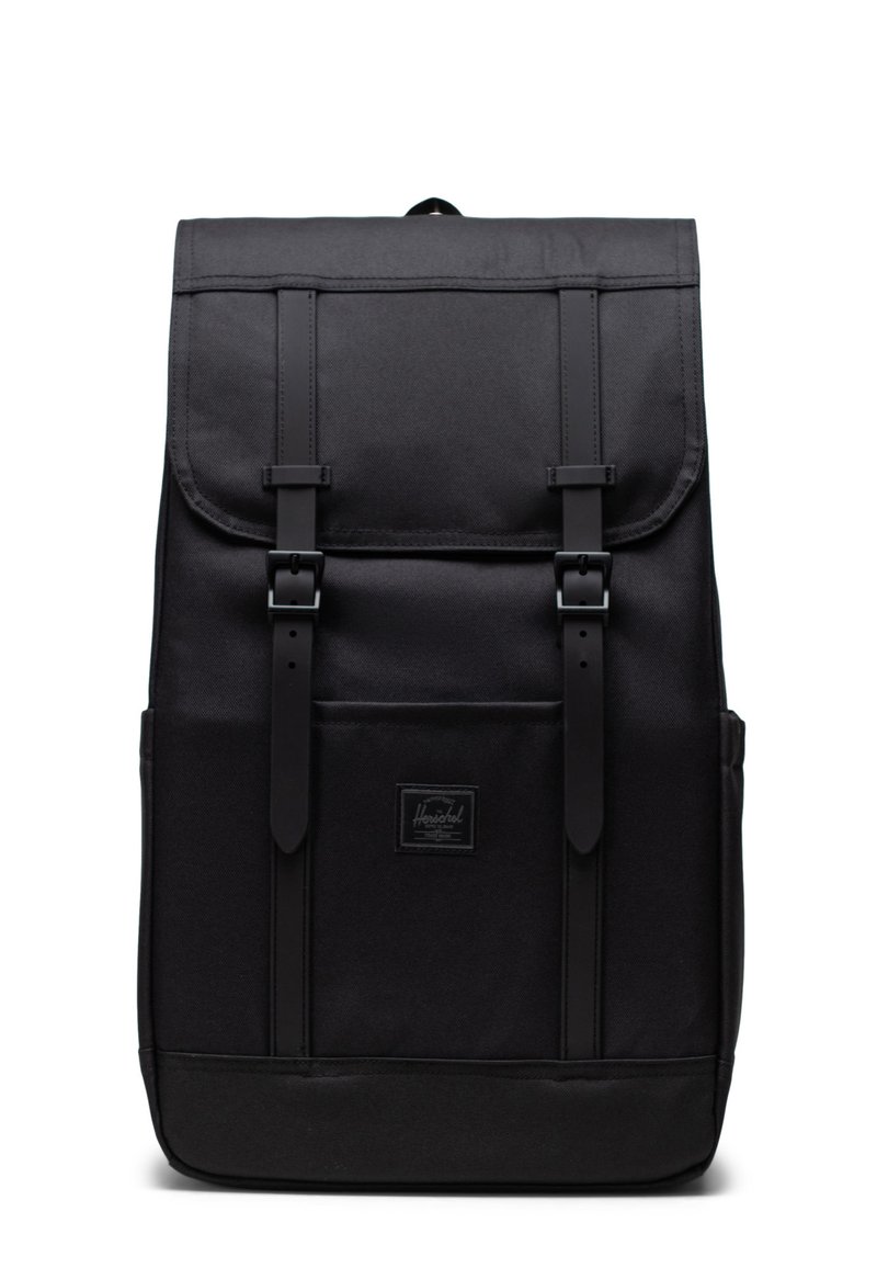 Zaino in canvas nero con pattina, due cinturini frontali, patch con logo e forma strutturata. Presenta una maniglia superiore e tasche laterali.