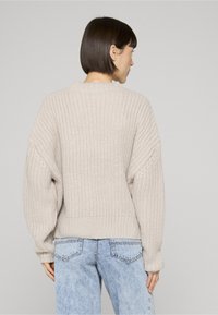 Frau mit kurzen dunklen Haaren, die einen beigefarbenen, grob gestrickten Pullover und hellblaue Jeans trägt, von hinten vor einem einfarbigen Hintergrund gezeigt.