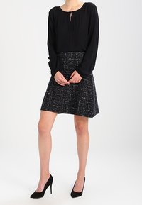 Schwarze Langarmbluse mit Schlüsselloch-Ausschnitt und plissierter schwarz-weißer Strukturrock. Kombiniert mit schwarzen High Heels.