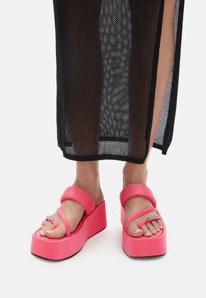 Piedi che indossano sandali rosa acceso con zeppa con due cinturini, abbinati a una gonna nera in rete che mostra gambe nude.