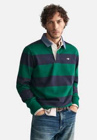 Groen en marineblauw gestreepte langslijf polo met een denim kraag, met eronder een wit overhemd en een logo op de borst.