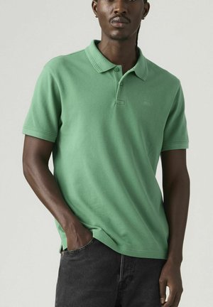 Mand iført grøn kortærmet polo-shirt og sorte jeans, stående med den ene hånd i lommen mod en neutral baggrund.