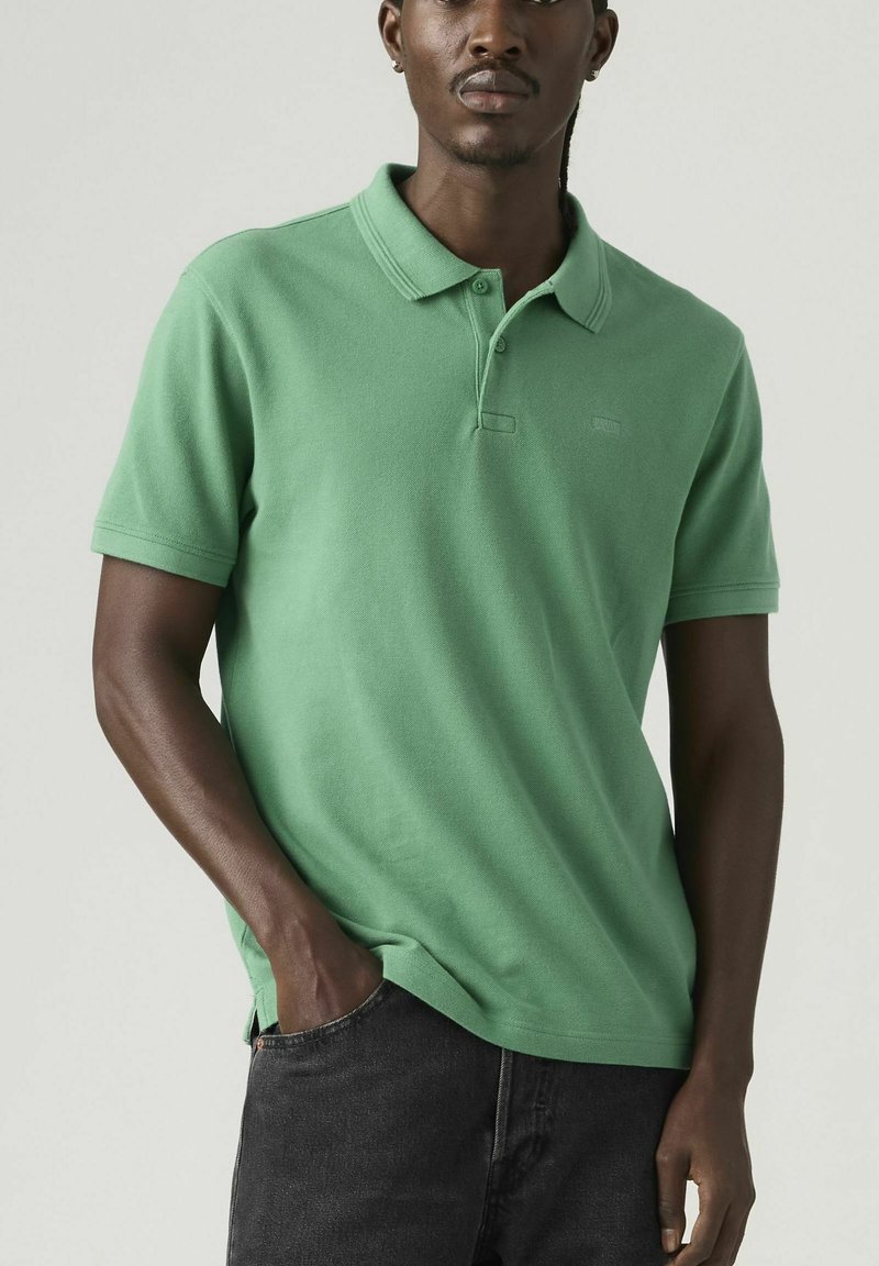 Hombre con polo verde de manga corta y vaqueros negros, de pie con una mano en el bolsillo contra un fondo neutro.