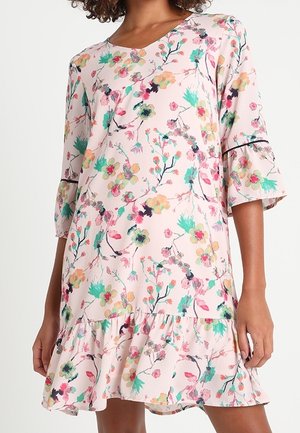 Kvinde iført en lys lyserød blomsterkjole med 3/4 klokkeærmer og en flæset kant med grønne, gule og lyserøde blomster.