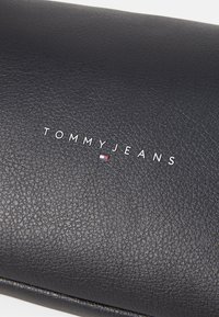 Crni kožni novčanik s teksturiranom površinom. "TOMMYJEANS" je uvučen u bijelo, uz malu obojenu zastavicu.