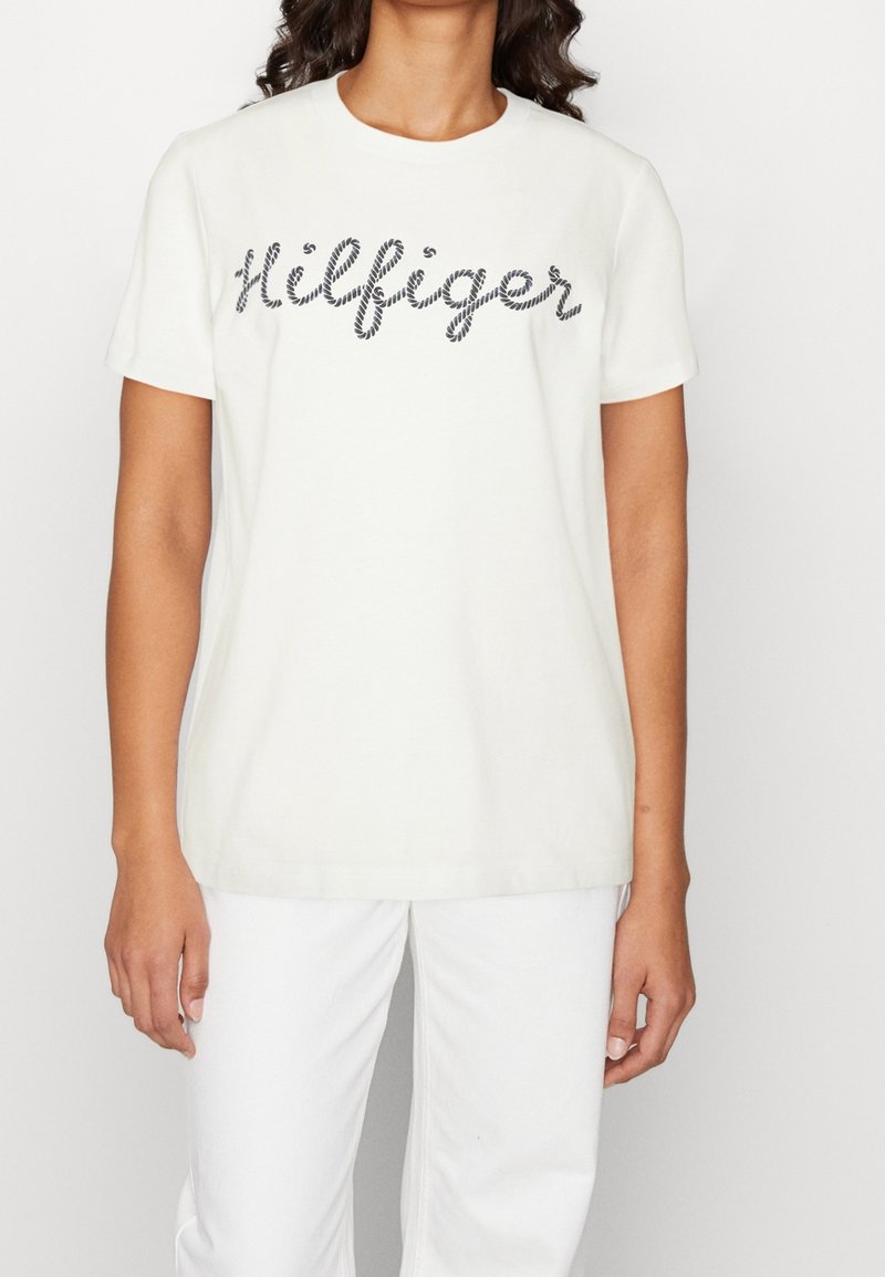 Femme portant une chemise blanche à manches courtes avec le texte "Hilfiger" en style corde noire et un pantalon blanc, debout devant un fond uni.