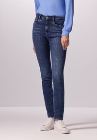 Vaqueros skinny azul oscuro de denim, con un clásico diseño de cinco bolsillos y cierre frontal de botón, combinados con zapatillas blancas.