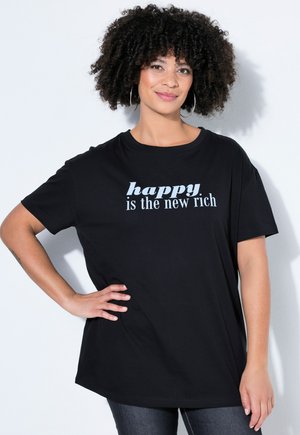 Mujer con cabello rizado y pendientes de aro que lleva una camiseta negra con la frase "feliz es el nuevo rico", de pie con una mano en la cadera.