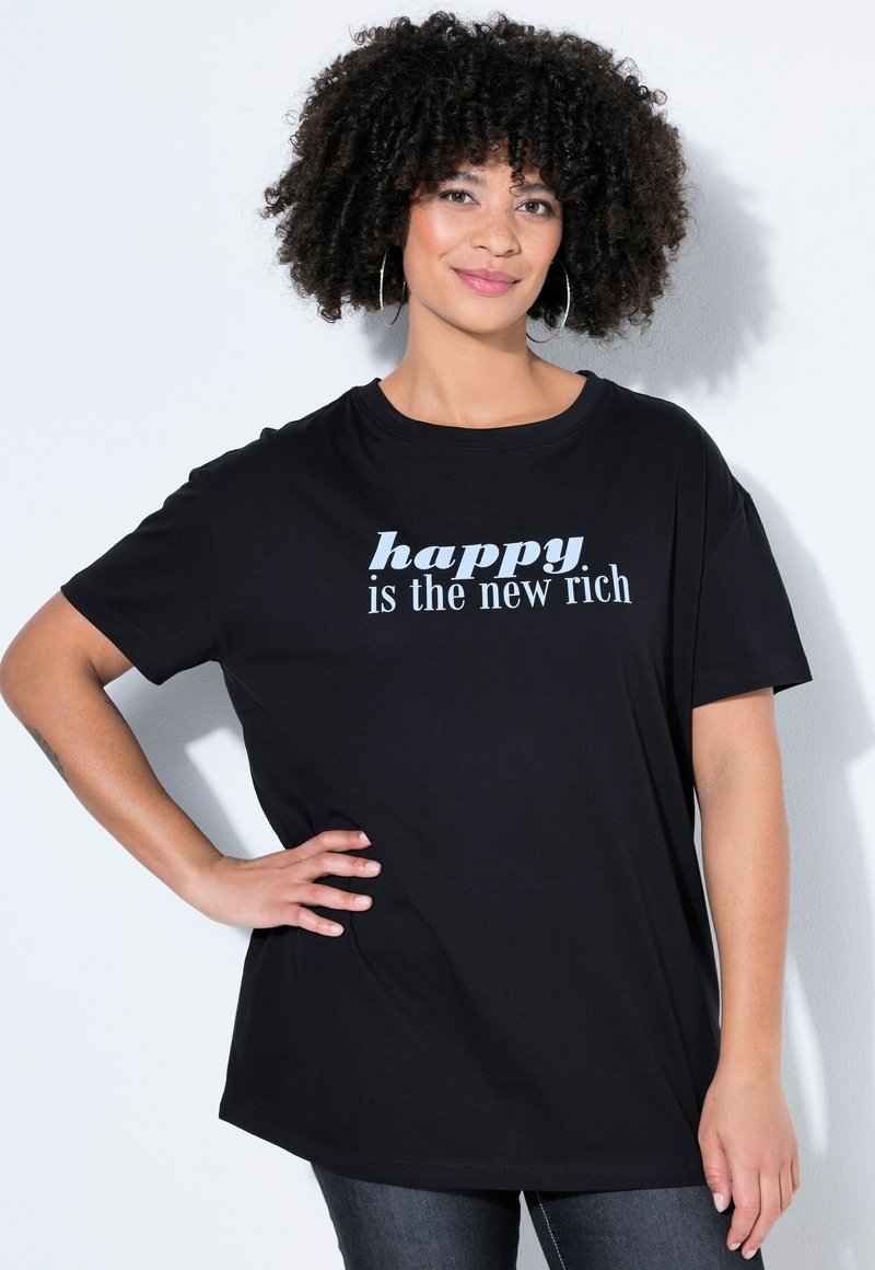 Kobieta o kręconych włosach i kolczykach kołach, ubrana w czarną koszulę z napisem "szczęście jest nowym bogactwem", stoi z jedną ręką na biodrze.