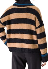 Personne portant un pull duveteux avec de larges rayures horizontales beige et noires et une bordure bleue au col et aux poignets, associé à un pantalon noir.