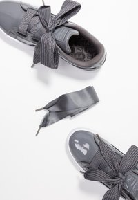 Paire de baskets Puma grises brillantes avec des lacets gris épais noués en boucle, à côté d'un long lacet séparé en satin gris sur fond blanc.