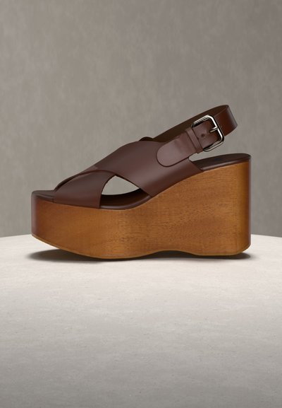 Sandale compensée en cuir marron avec talon compensé en bois et bride de cheville réglable avec boucle en argent sur fond neutre.