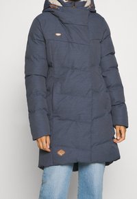 Marinblå puffjacka med huva, med en strukturerad tyg, horisontell sömnad och en brun logotyp patch på nedre fållen.