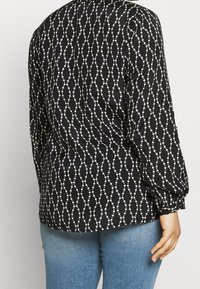 Sort bluse med et hvidt cirkulært og bølget mønster, med lange ærmer med samlede manchetter, fremstillet af et glat stof.