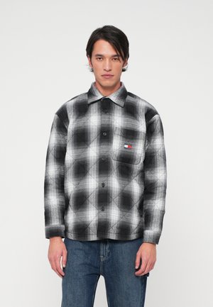 Camisa de flanela acolchoada em padrão xadrez preto e branco, com frente de botões e um bolso no peito com um patch de logo. Jeans de ganga completam o visual.