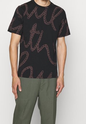 T-shirt noir à manches courtes avec motif abstrait de serpent marron, porté avec un pantalon vert olive, visible du cou au milieu des cuisses.