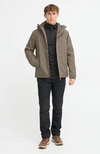 Chaqueta verde oliva con cremallera en la parte delantera, capucha y dos bolsillos laterales, llevada sobre una camiseta negra de cuello alto, combinada con pantalones negros y zapatos marrones.