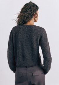 Kvinde med bølget mørkt hår iført en mørk, blød sweater og mørke bukser, stående med hænderne i lommerne, vendt væk mod en ensfarvet baggrund.