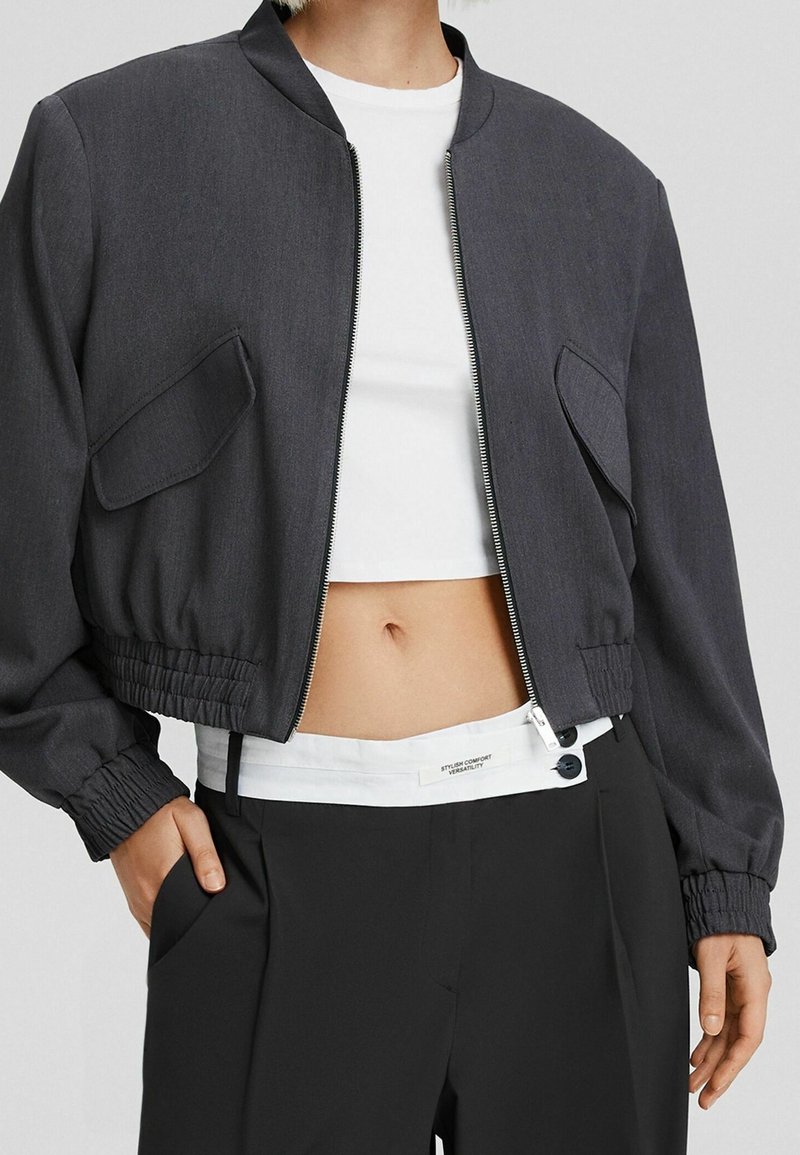 Grå bomberjacka med en strukturerad design, ribbad mudd och två framfickor. Bärs över en vit crop top och ihop med svarta byxor.