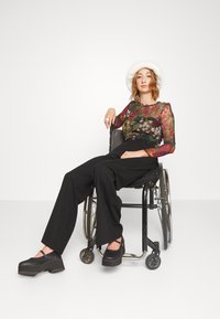 Svart rullestol med sølvfargede detaljer. Modellen har på seg en gjennomsiktig blomstret topp og vide svarte bukser, kombinert med svarte platå sko og en hvit hatt.