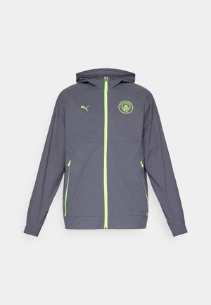 Giacca leggera grigia con cappuccio, zip verde e logo del Manchester City. Presenta polsini elastici e tasche laterali.