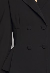 Černá dvouřadá bunda s velkými knoflíky, na míru střižená a s peplum detailem. Vyrobena z hladké látky se strukturovaným designem a ostrými klopami.