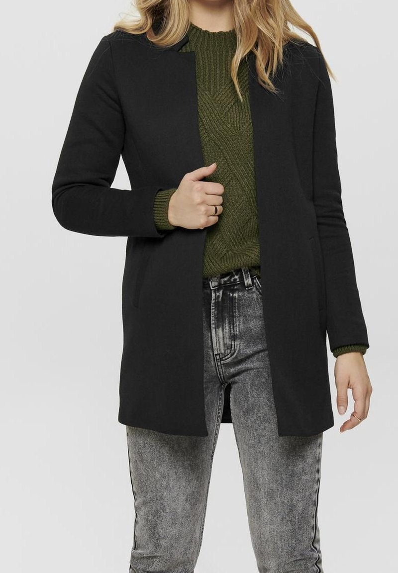 Manteau noir sur mesure à coupe droite, tissu léger, sans fermetures visibles, porté sur un pull en maille vert et un jean gris.