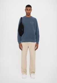 Hanorac albastru cu textul "Doza zilnică de denim", pantaloni largi bej și o geantă neagră tip crossbody. Adidași albi completează ținuta.