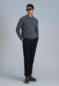 Pull en maille côtelée grise avec col rond, associé à un pantalon ajusté bleu marine foncé et des chaussures en cuir à lacets marron, mannequin portant des lunettes de soleil noires.