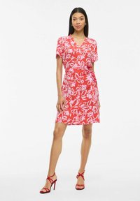 Abito floreale rosso con maniche corte, scollatura a V, vita strappata e lunghezza al ginocchio. Presenta motivi floreali rosa ed è abbinato a sandali rossi con cinturini.