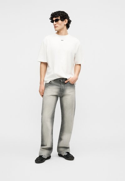 Άνδρας που φοράει μαύρα γυαλιά ηλίου, λευκή φαρδιά μπλούζα, ξεθωριασμένο γκρι τζιν ίσιας γραμμής και μαύρα αθλητικά με λευκές ρίγες, στέκεται με το χέρι στην τσέπη.