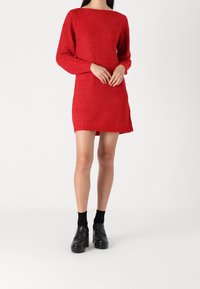 Una donna indossa un abito rosso a maglia, con maniche lunghe e scollatura a barca, abbinato a scarpe con platform nere e calzini alla caviglia.