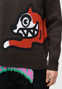 Πουλόβερ με μεγάλο καρτουνίστικο πορτοκαλί και λευκό πρόσωπο αλεπούς με σπειροειδή μάτι και αιχμηρά δόντια σε σκούρο καφέ ύφασμα, φορεμένο με μαύρο παντελόνι.