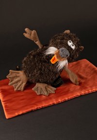 Plush dark brown creature with light brown feet, a gray snout, and orange accents, resting on a smooth orange fabric surface.