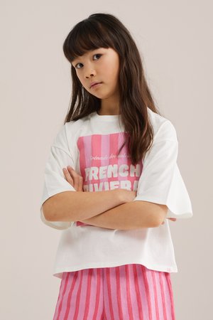 Młoda dziewczyna z długimi ciemnymi włosami, ubrana w biały T-shirt z różowym napisem „French Riviera” oraz różowe spodnie w paski, z założonymi skrzyżowanymi ramionami.