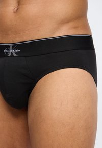 Κοντινό πλάνο ενός ατόμου που φοράει μαύρα εσώρουχα Calvin Klein, εμφανίζοντας το λογότυπο στη μέση και την περιοχή του άνω μηρού.