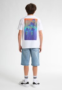 Hvid t-shirt med et farverigt grafisk mønster af abstrakte figurer på ryggen. Parret med lyseblå denimshorts og hvide sneakers med sorte detaljer.