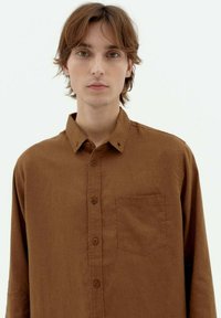 Brown button-up shirt made of textured fabric, featuring a chest pocket and a classic collar. The design is simple and fitted.
